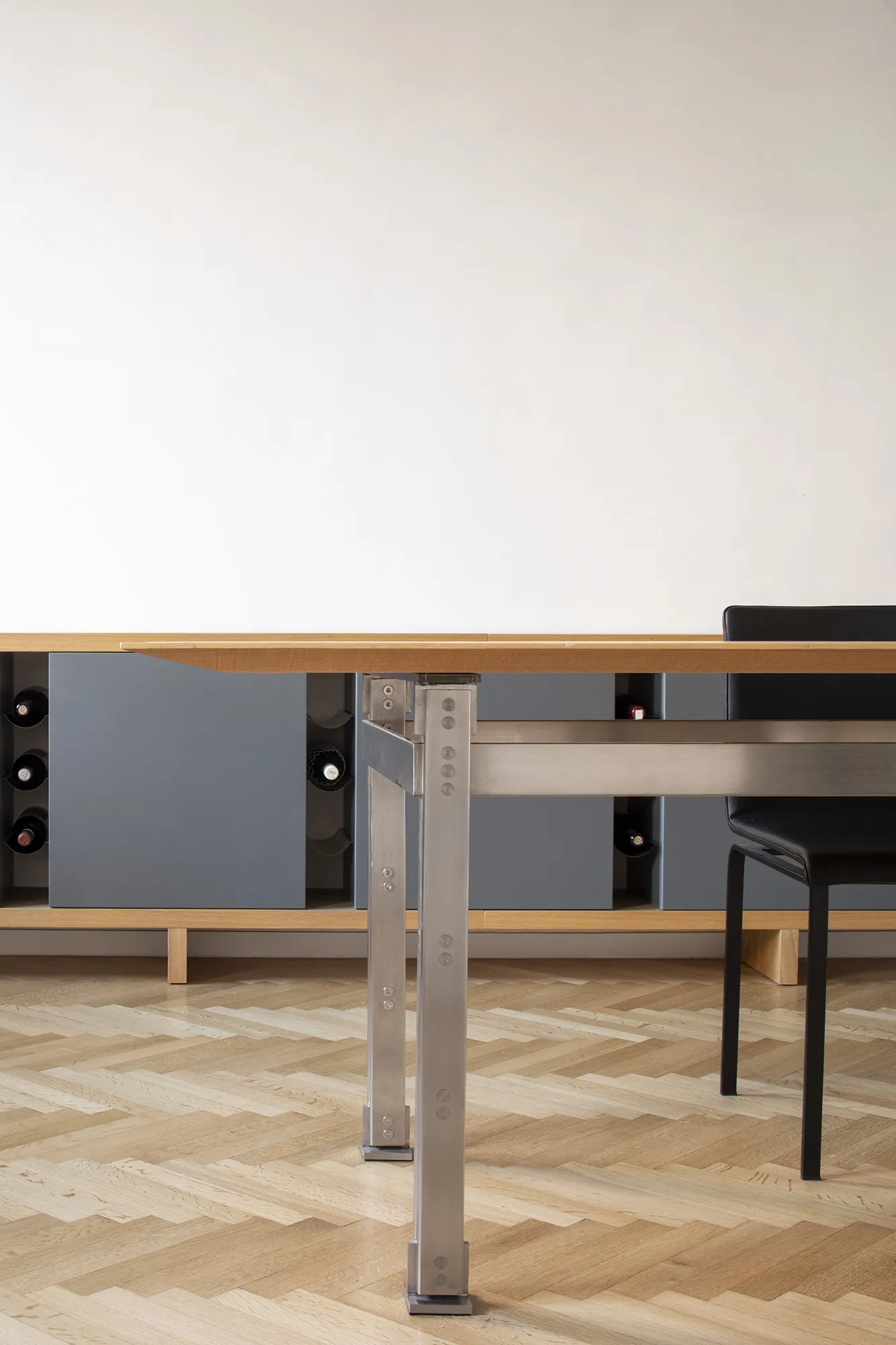 Vista frontale di un tavolo di design in legno e acciaio davanti a una madia grigia con vani porta bottiglie in un interno di design.