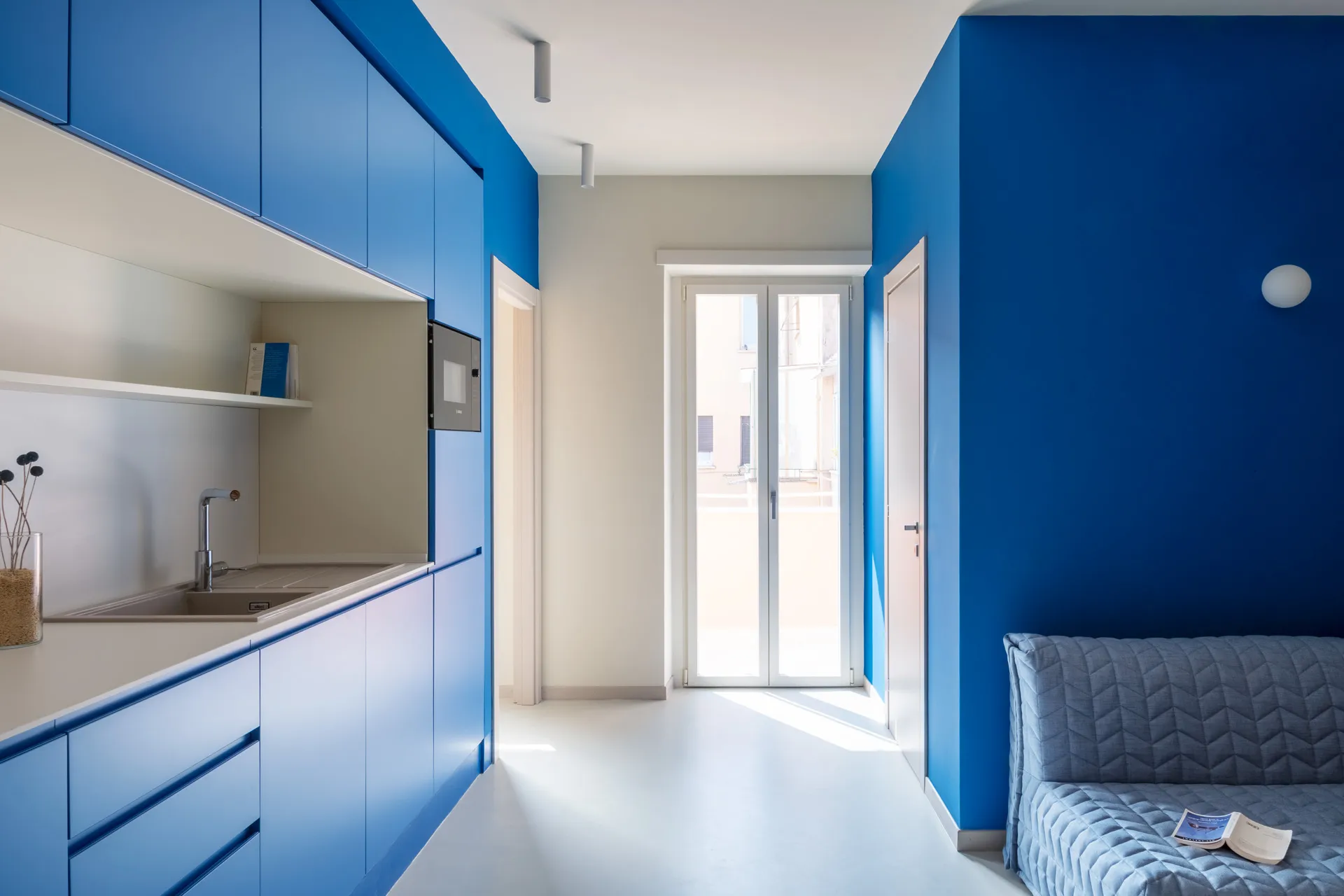 Arredo open space per casa vacanze con cucina blu monoblocco e divano coordinato su pavimento bianco.