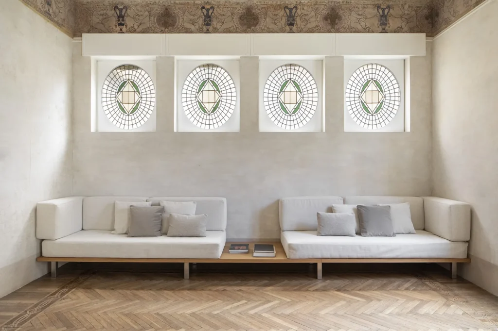 Salone moderno in palazzo storico con quattro finestre a oblò decorate di epoca liberty , fregio affrescato e divani minimalisti su misura su pedana in legno.