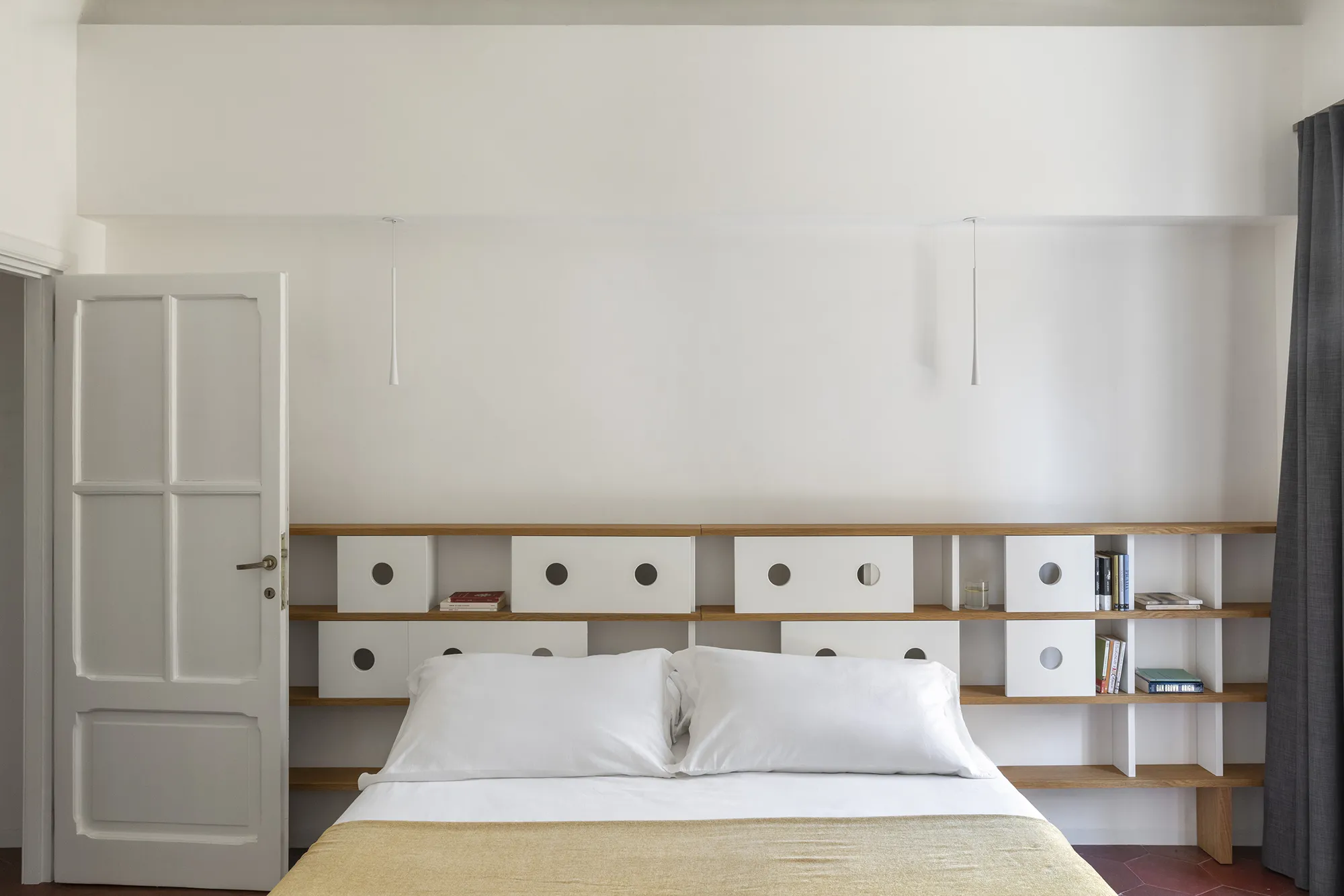 Testiera del letto moderna integrata in una scaffalatura in legno con moduli bianchi a oblò e lampade a sospensione minimali.
