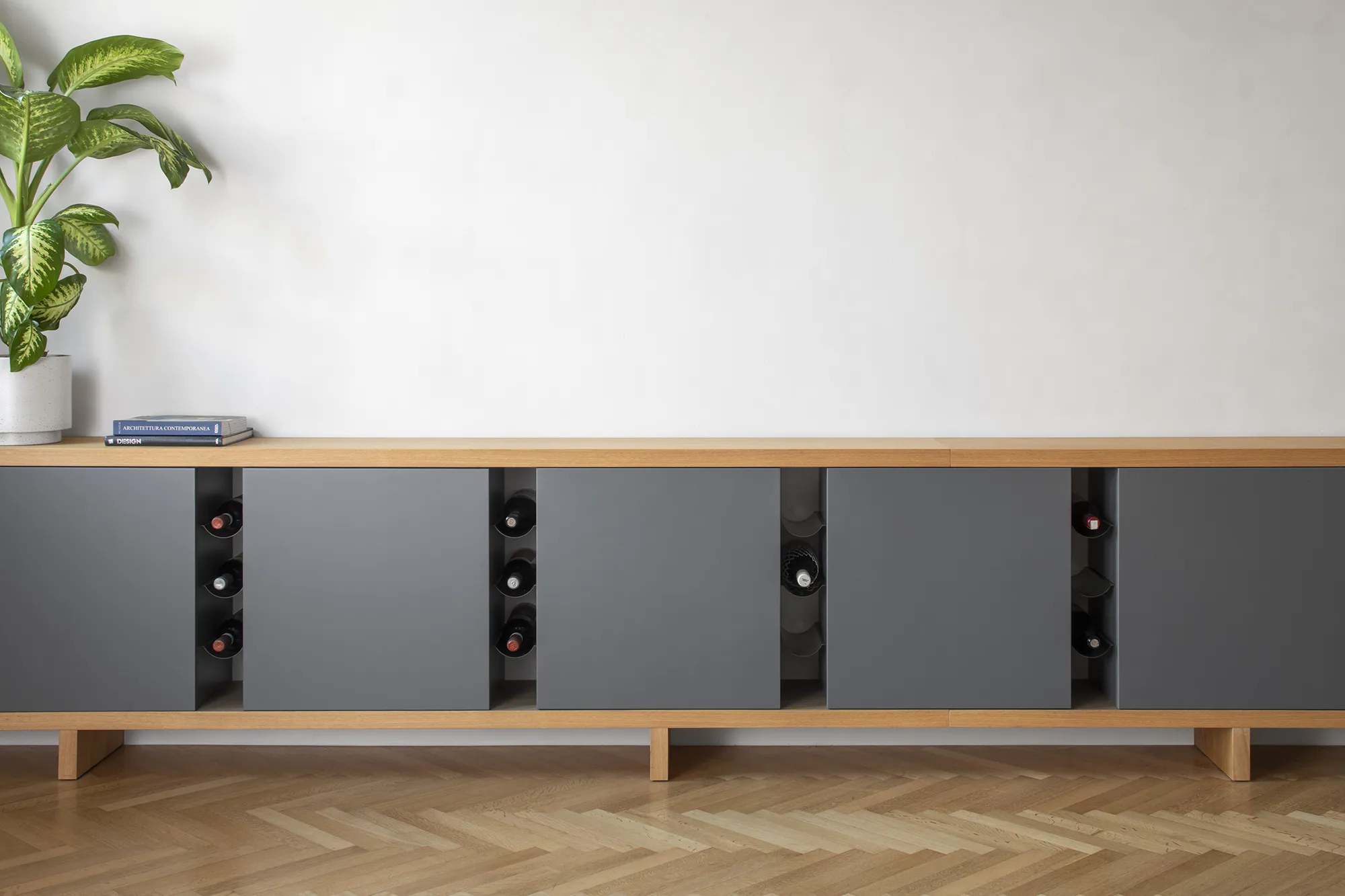 Madia moderna grigio antracite con vani porta bottiglie integrati, piano in rovere e pianta decorativa in un interno di design.
